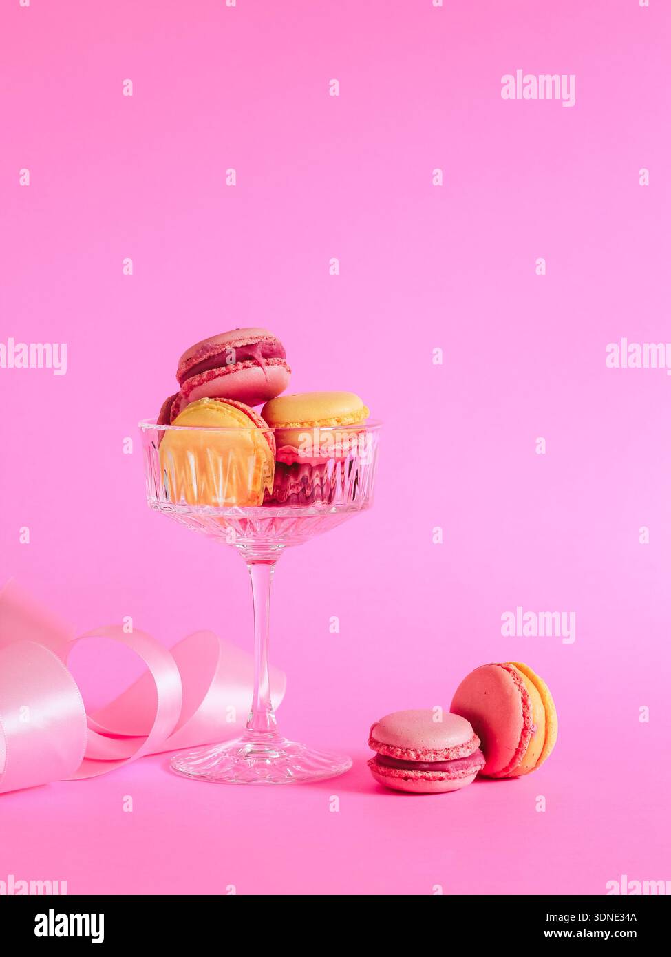 Herzlichen Glückwunsch zum Valentinstag. Rosafarbene und gelbe Macarons in einem Champagnerglas und rosafarbenes Band auf rosa Hintergrund. Stockfoto