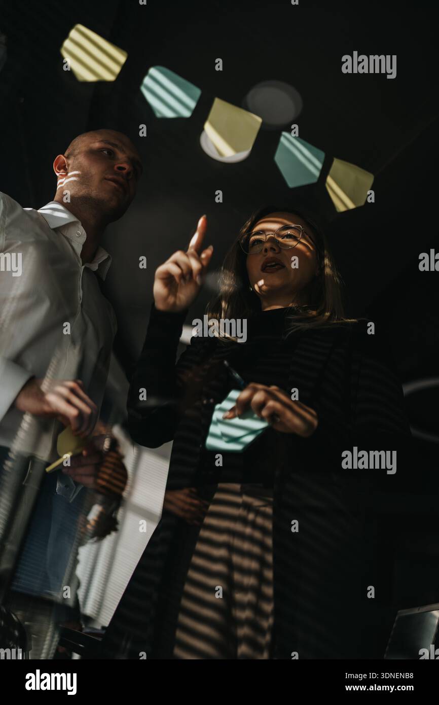Zwei Kollegen ziehen ein Brainstorming unter dekorativen Bannern durch und platzieren während einer gemeinsamen Bürositzung Haftnotizen Stockfoto