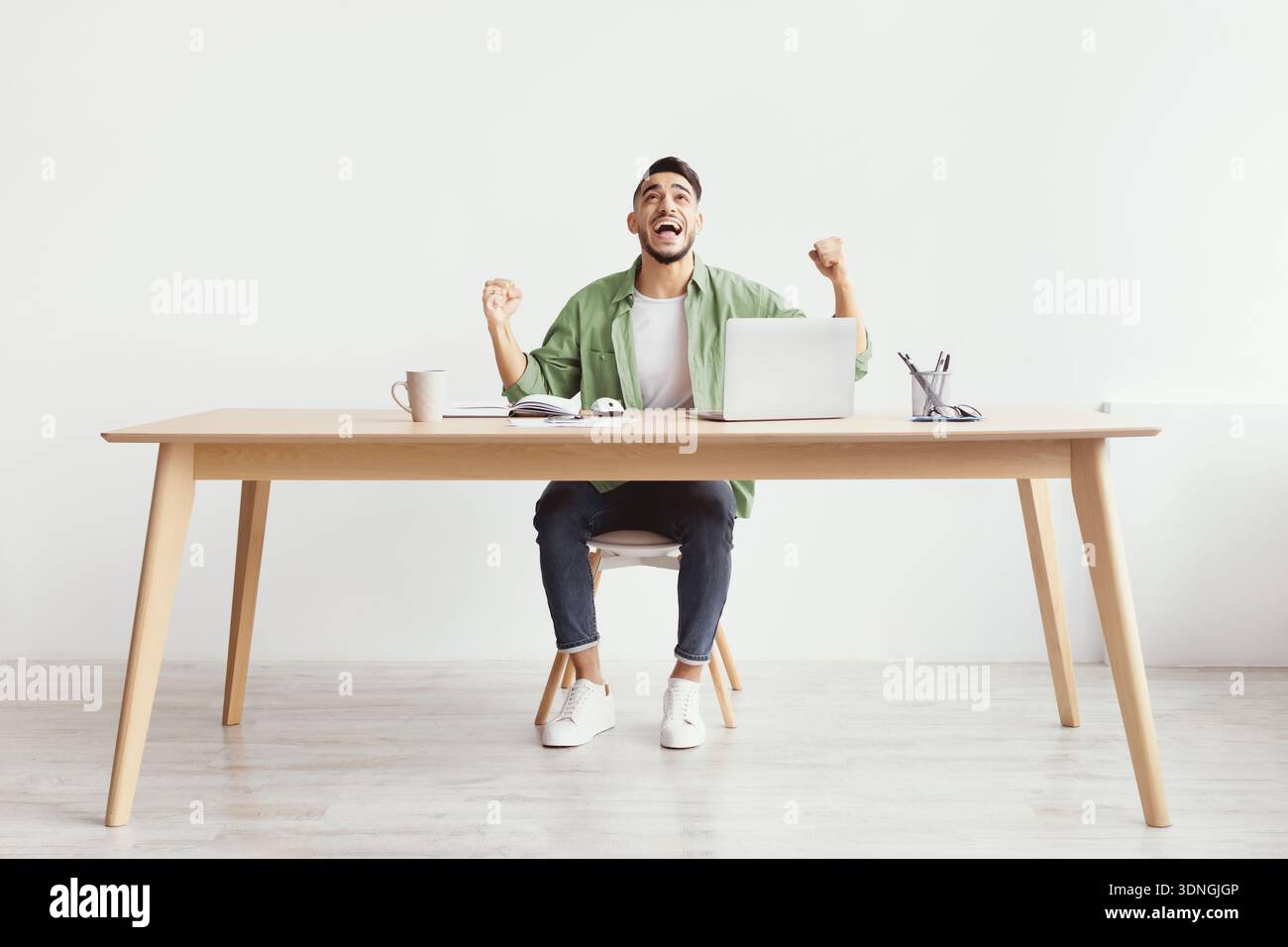 Mann feiert Erfolg, während er an einem unordentlichen Schreibtisch im Home Office sitzt Stockfoto
