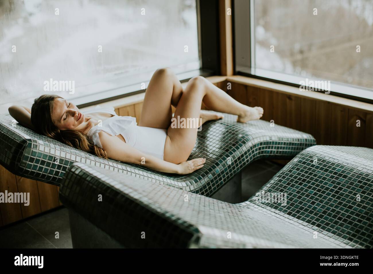 Eine Frau liegt auf einem warmen Steinbett in einem Spa. Sie genießt die warme Oberfläche und die Aussicht nach draußen. Die Umgebung ist gemütlich und einladend, perfekt zum Entspannen Stockfoto