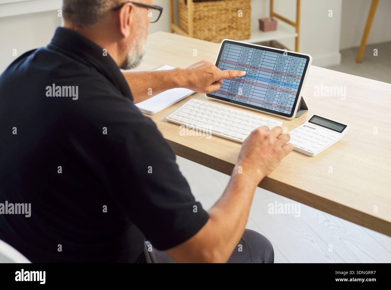 Senior-Mann analysiert Tabellenkalkulationen auf Tablets mit Rechner im aufgeräumten Home Office Stockfoto