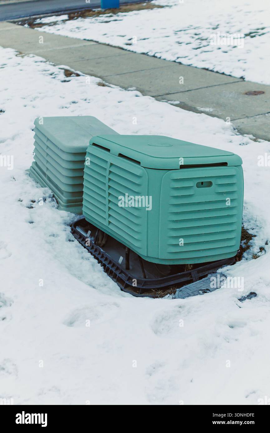 Zwei grüne Kompostbehälter stehen auf einem Schneefleck in einer Nachbarschaft. Der Boden ist mit weißem Schnee bedeckt, was Anzeichen von Winter zeigt. In der Nähe befindet sich ein Bürgersteig. Stockfoto