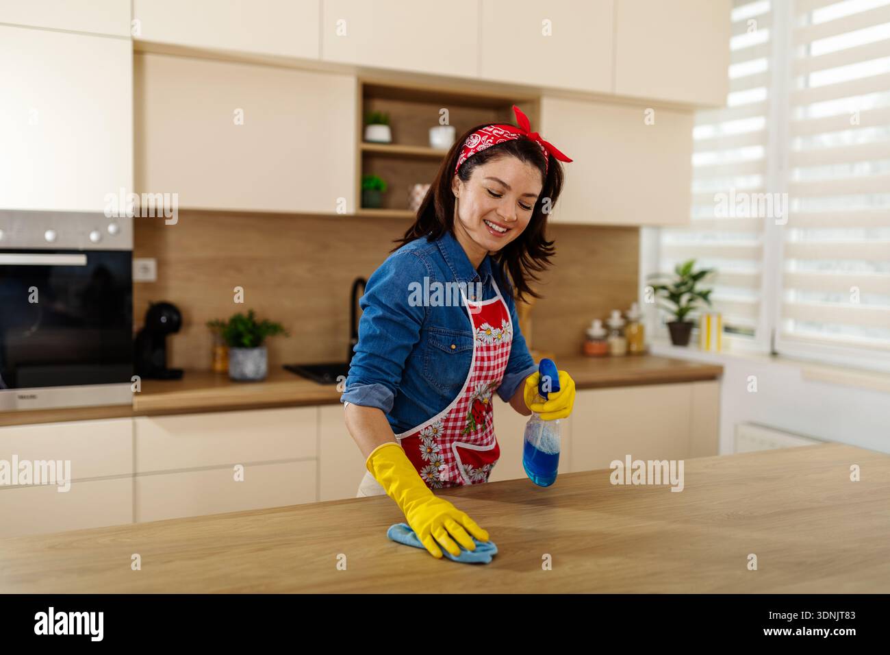Eine Frau in gelben Handschuhen benutzt eine Sprühflasche und ein Tuch, um eine Küchenoberfläche zu reinigen. Sie konzentriert sich auf ihre Aufgabe in einer hellen Küche Stockfoto