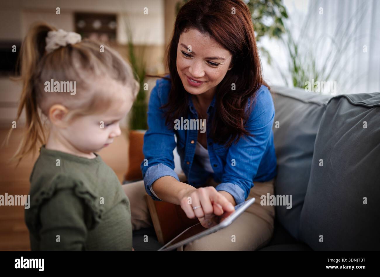 Eine Mutter zeigt ihrer Tochter eine Tafel, während sie auf einer Couch sitzt. Sie befinden sich in einem hellen Wohnzimmer mit Pflanzen und einer warmen Atmosphäre. Stockfoto