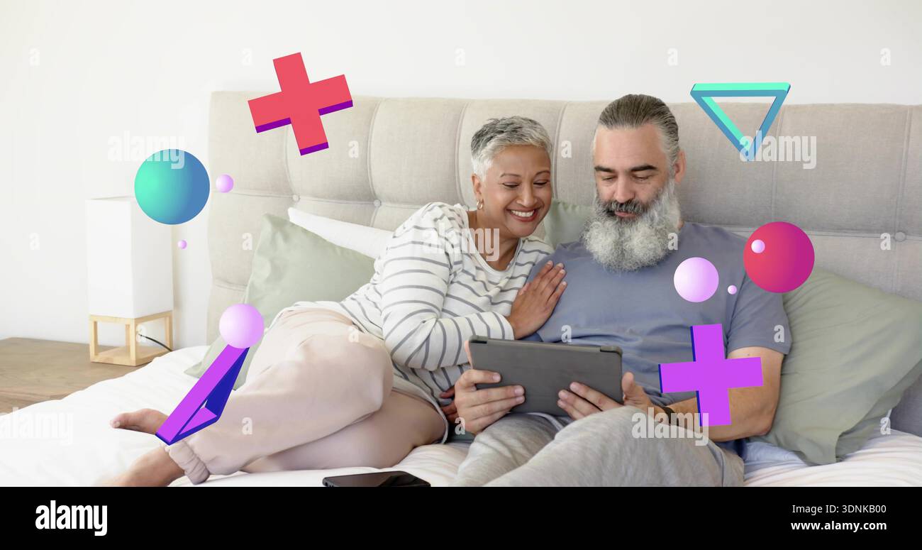 Liegendes reifes Paar, das sich ein Tablet auf dem Bett im Schlafzimmer teilt, Mann mit Bart, Frau in gestreiftem Oberteil Stockfoto