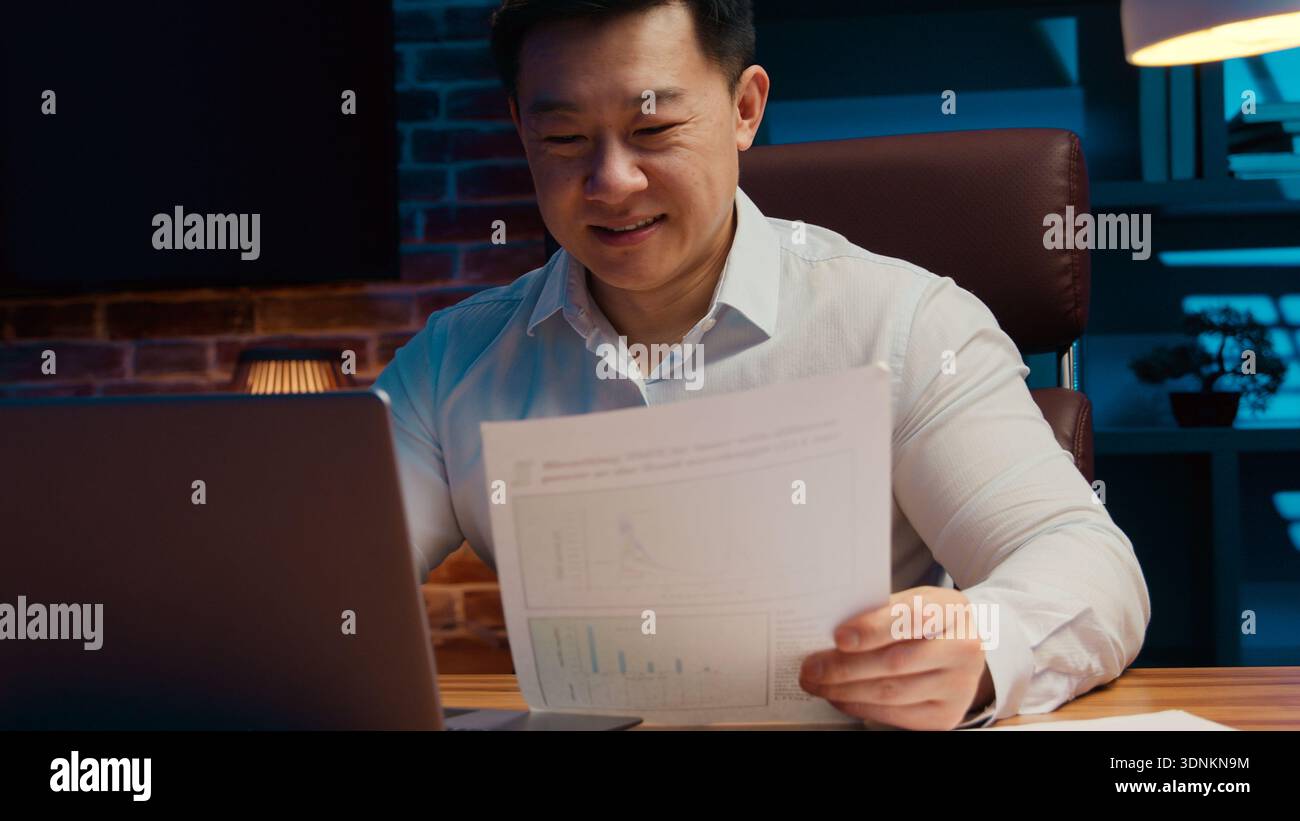 Lächelnder koreanischer Geschäftsmann mittleren Alters Schreib Laptop Rezensionspapiere Unternehmen Gute Finanzergebnisse im Nachtbüro chinesischer Geschäftsmann Lächeln Stockfoto