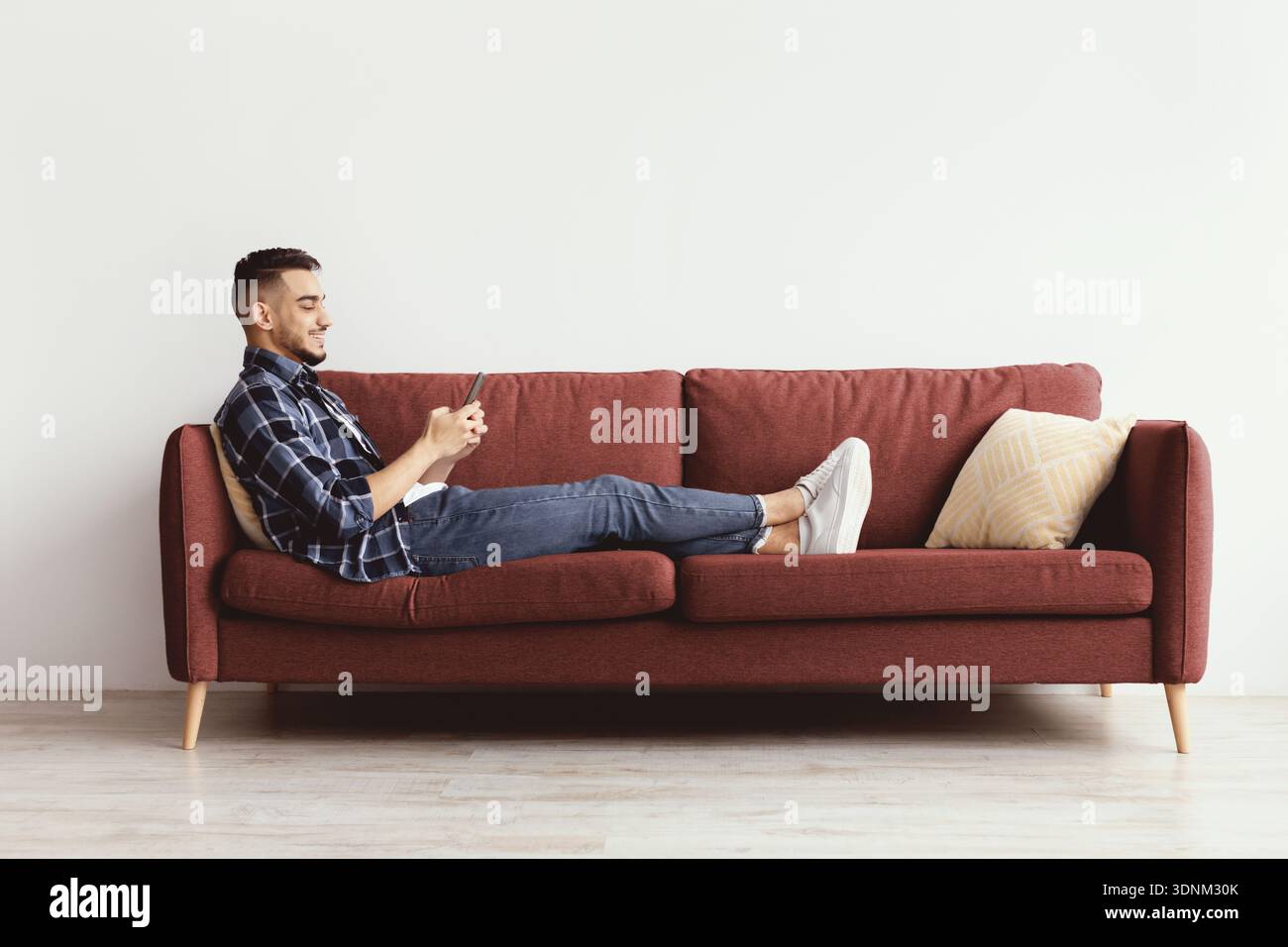 Mann, der auf dem roten Sofa sitzt und das Smartphone in einem einfachen Innenraum benutzt Stockfoto