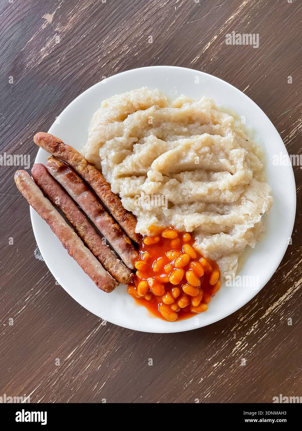 Die Ansicht von oben zeigt einen Teller mit Würstchen, Bohnen in Tomatensauce, Kartoffelpüree auf Holztisch. Herzhafte hausgemachte Mahlzeit mit klassischer Komfortspeise Stockfoto