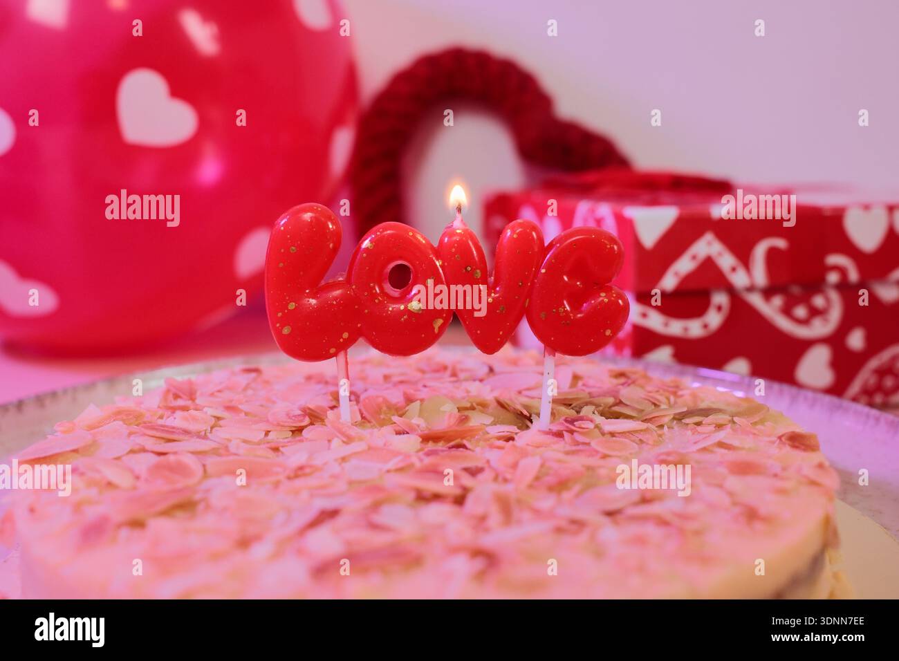 Romantischer Kuchen mit Mandelsplittern und einer brennenden roten Liebeskerze, umgeben von Herzdekorationen und Geschenkpapier. Festliches Dessertkonzept für Stockfoto