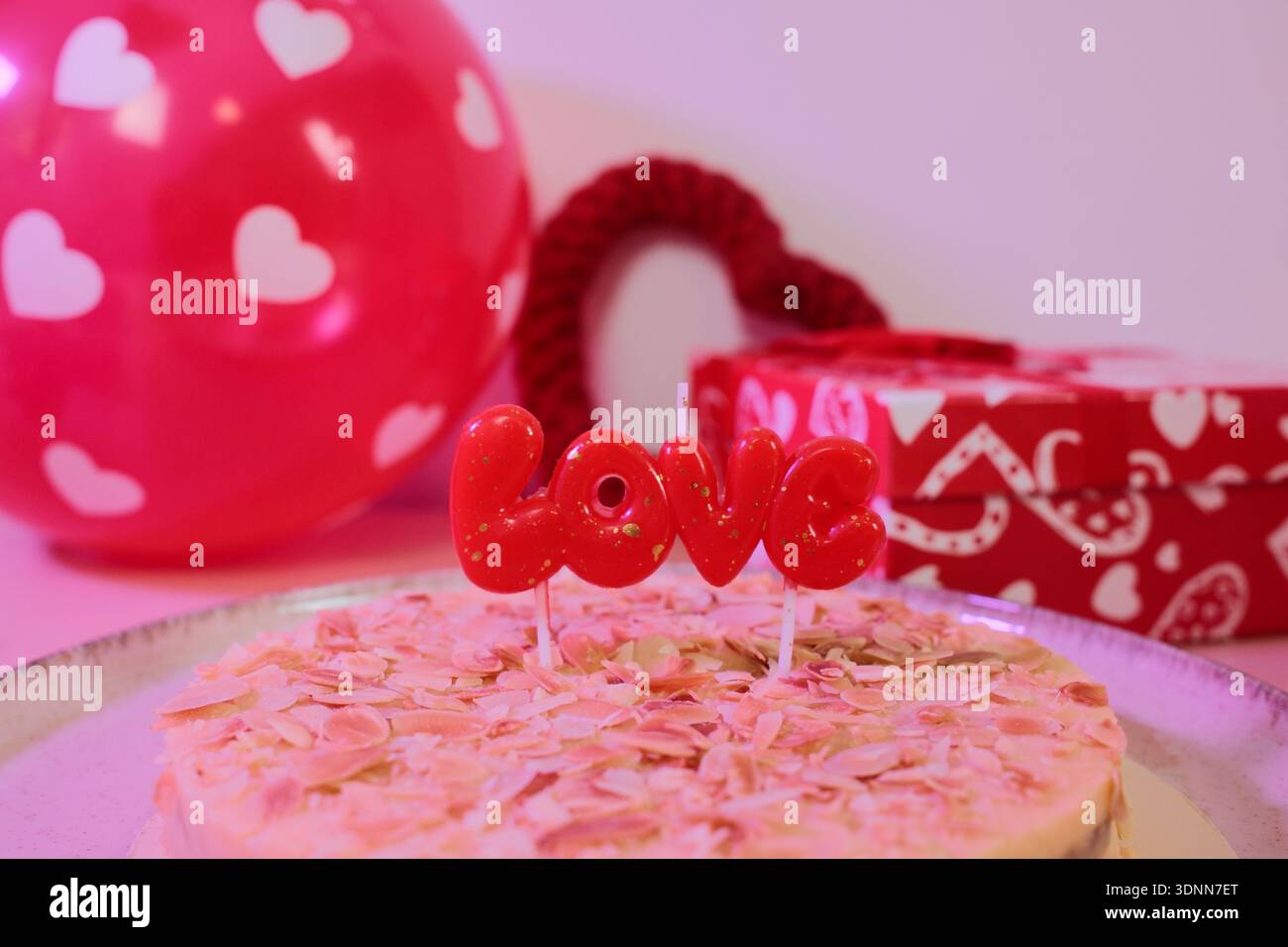 Romantischer Kuchen mit Mandelflocken und roter Liebeskerze, umgeben von Herzdekorationen und Geschenkbox. Festliches Dessertkonzept zum Valentinstag Stockfoto