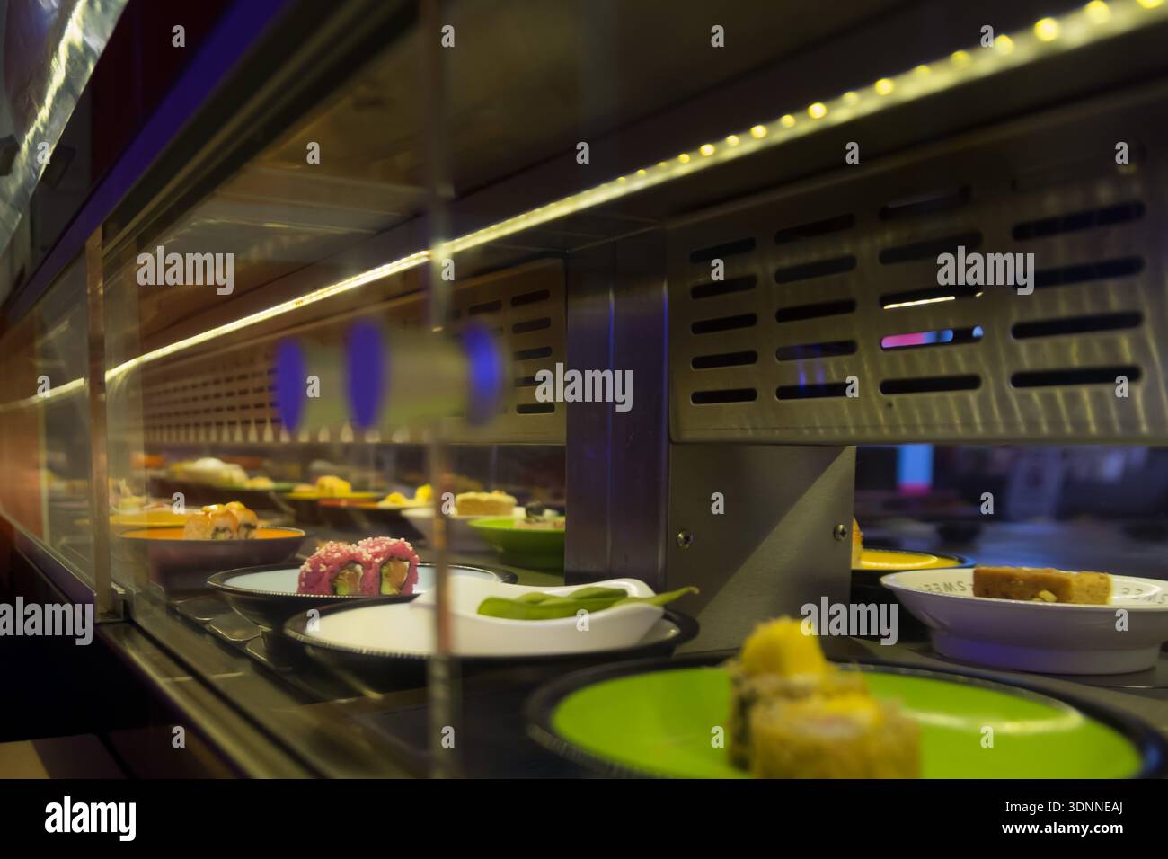 Eine perspektivische Nahansicht farbenfroher Platten mit verschiedenen Arten von Sushi, Rollen und Edamame, die sich auf einem rotierenden Förderband bewegen. Die Szene-Featur Stockfoto
