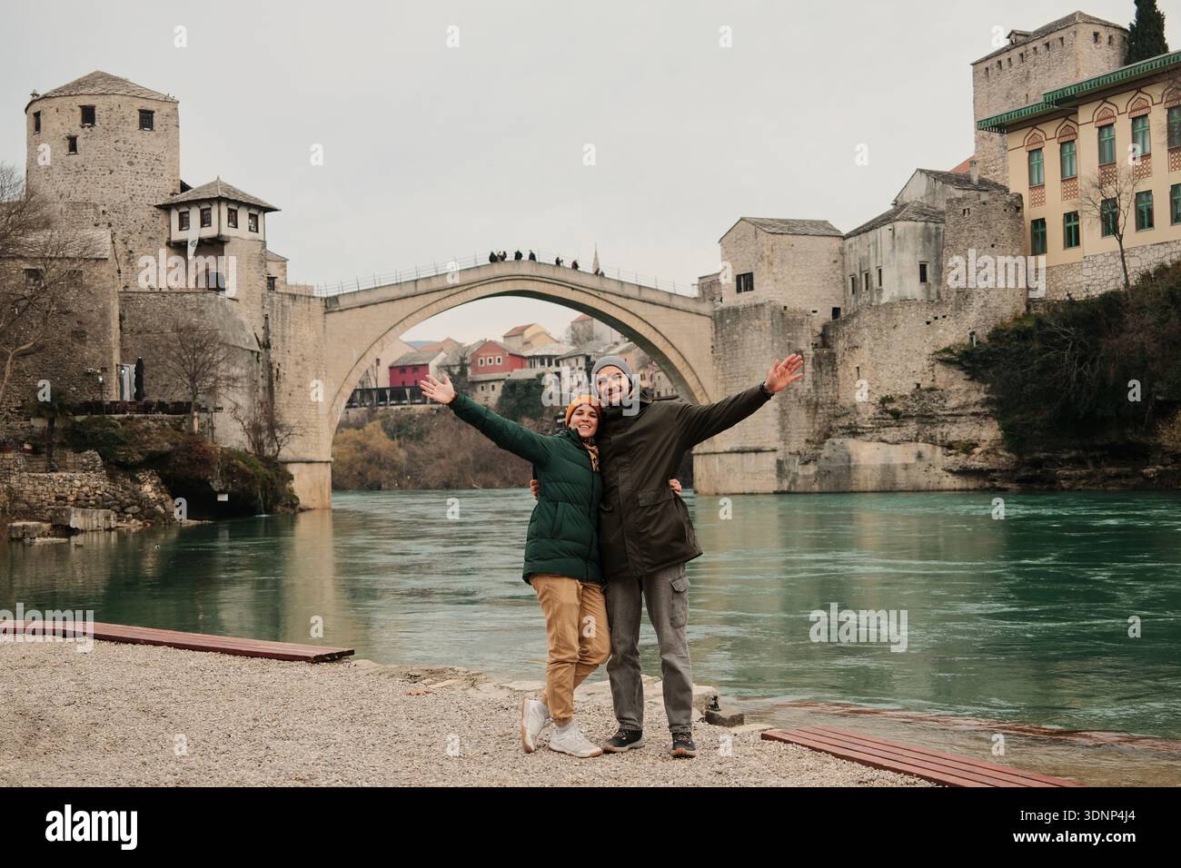 Lächelndes Paar mit offenen Armen am Fluss in der Nähe der historischen Brücke in Mostar. Reisefreiheit und gemeinsames Glücksgefühl, inspiriert von Stockfoto