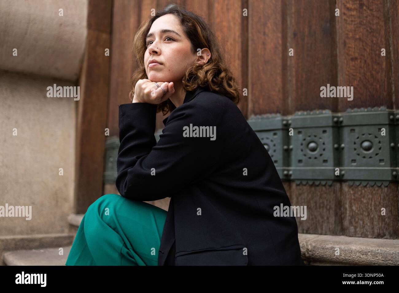Eine nachdenkliche Frau sitzt auf Steintreppen im Freien und legt ihr Kinn auf ihre Hand. Sie blickt nach oben, trägt einen schwarzen Blazer und eine grüne Hose gegen eine Te Stockfoto