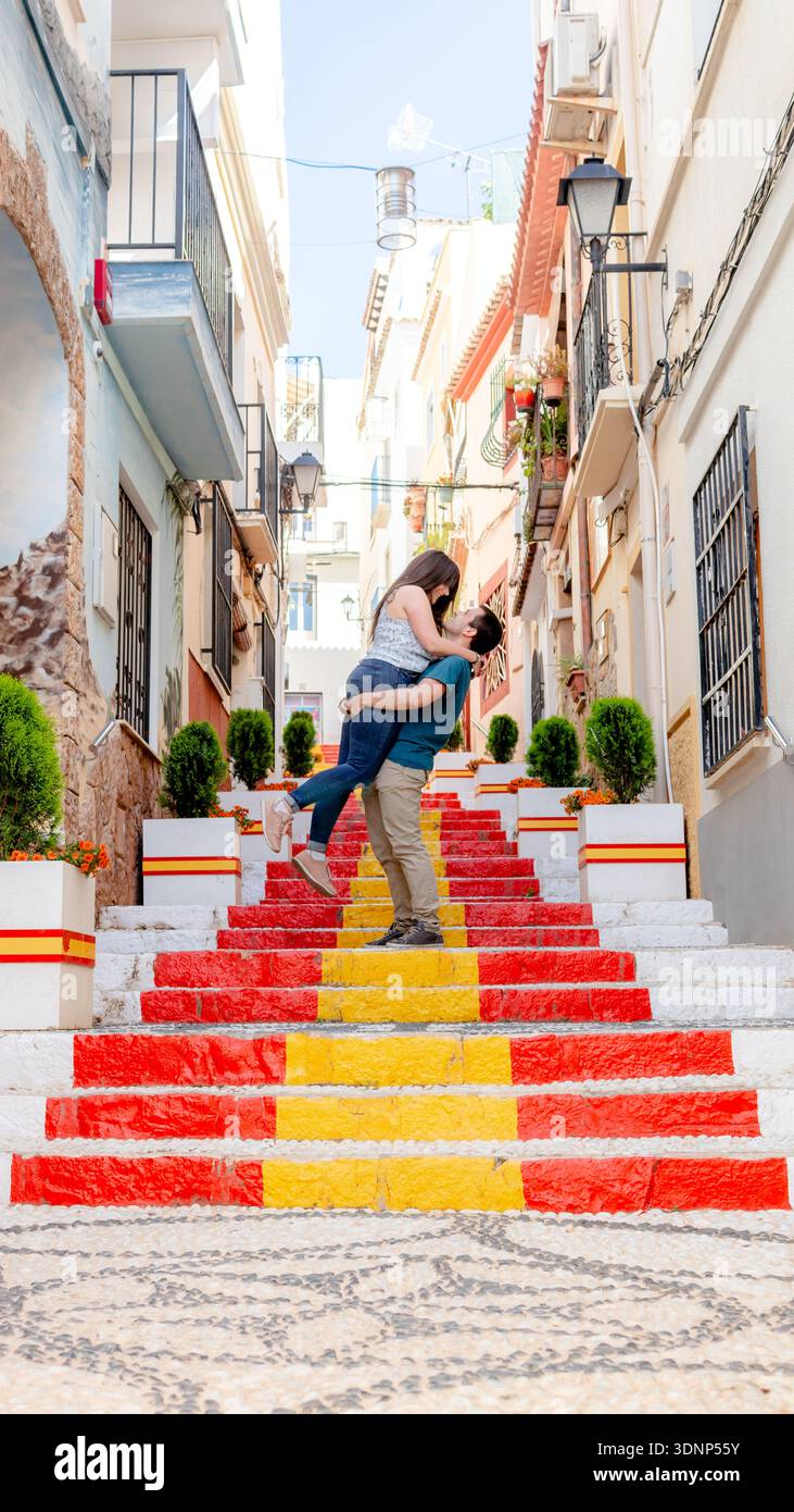 Ein Paar teilt einen romantischen Moment auf den farbenfrohen Stufen, die im Muster der spanischen Flagge gemalt sind. Das Hotel liegt in den malerischen Straßen von Alicante, Spanien, Stockfoto