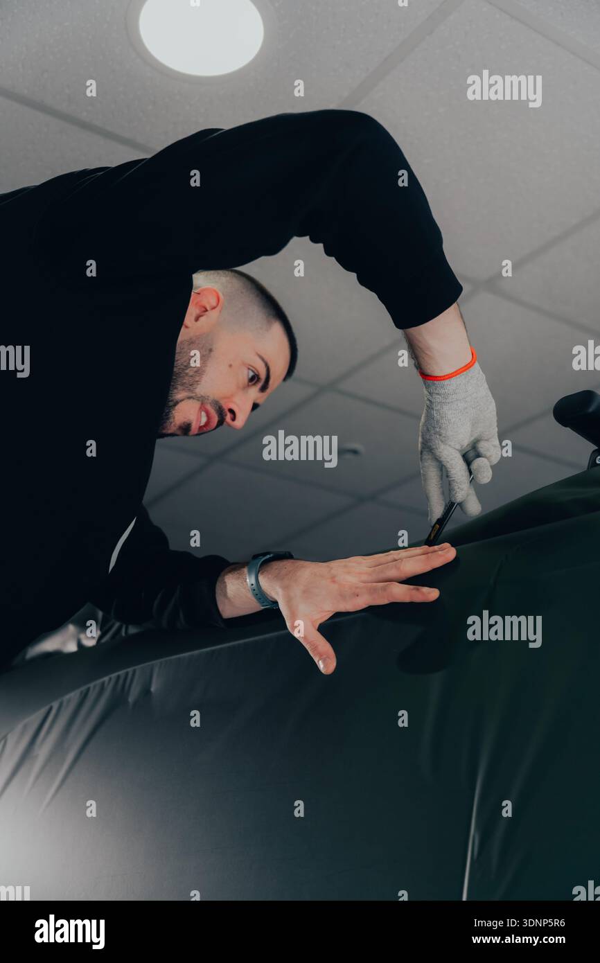 Ein professioneller Techniker in einer Autowerkstatt hüllt ein Fahrzeug fachmännisch mit Vinyl und zeigt die Kunst der individuellen Gestaltung des Fahrzeugs. Der Schwerpunkt liegt auf der Handwerkskunst Stockfoto