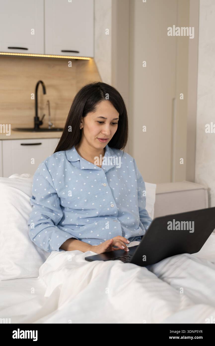 Eine Frau im bequemen Pyjama arbeitet von ihrem Bett aus mit einem Laptop und vermittelt so ein entspanntes und gemütliches Zuhause. Die Einstellung schlägt die Produktivität und vor Stockfoto
