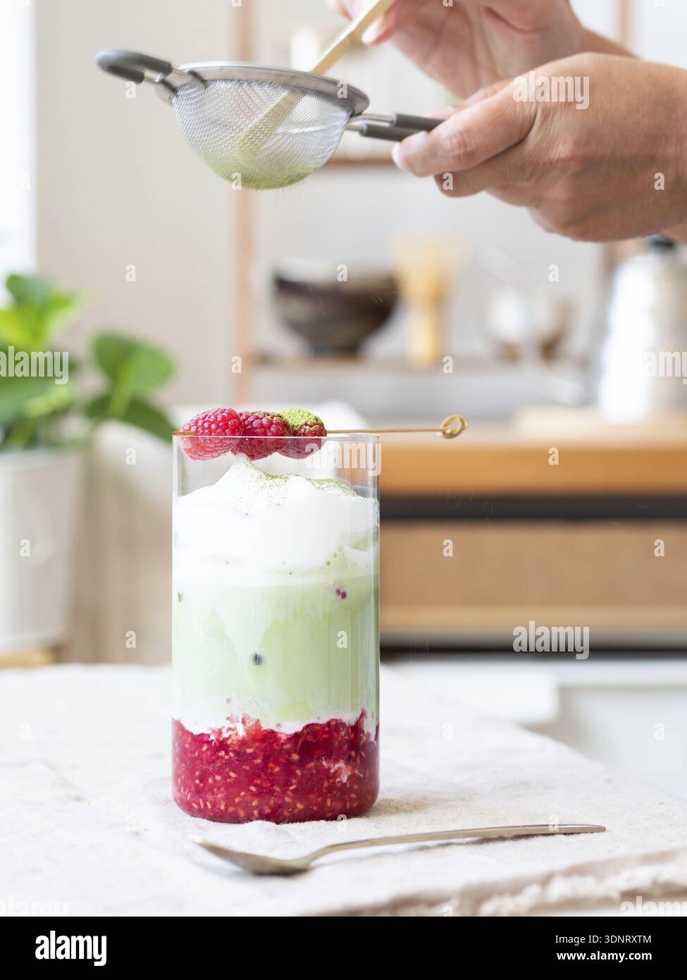 Person, die Grünteepulver über einem erfrischenden Eismatcha Latte mit Himbeeren siebt Stockfoto