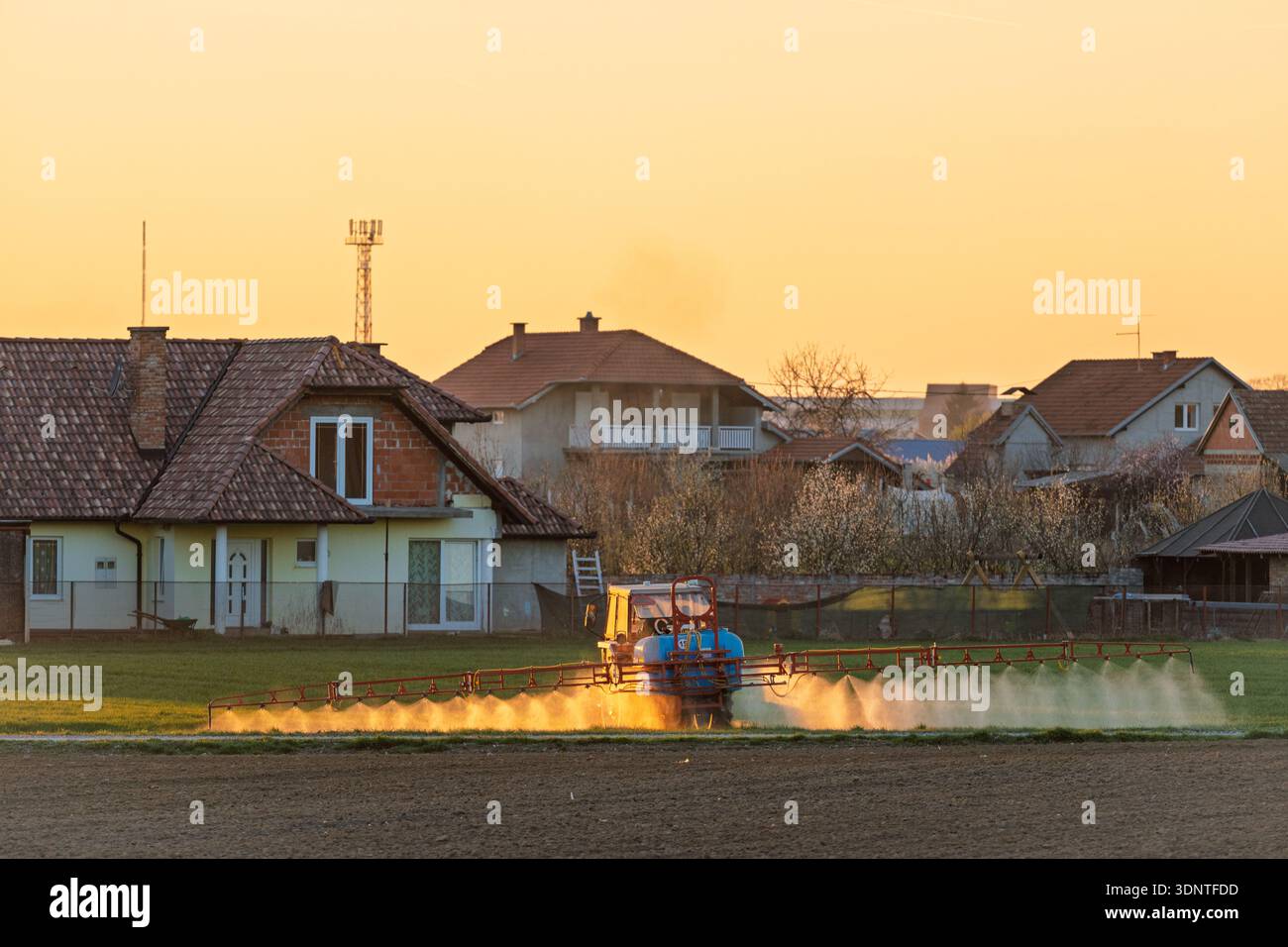 Eine ruhige ländliche Szene bei Sonnenuntergang verfügt über einen Sprinkler, der ein Feld vor Häusern und Gärten bewässert, was das ruhige und praktische Leben auf dem Land einfängt Stockfoto