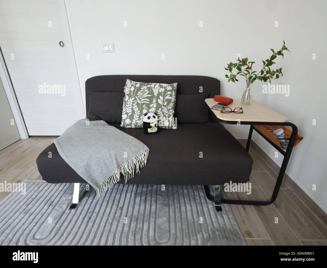 Kompaktes schwarzes ausklappbares Schlafsofa mit Überwurf und Kissen sowie Beistelltisch. Möbel für kleines Wohnzimmer. Helle Wände, grauer Teppich. Stockfoto