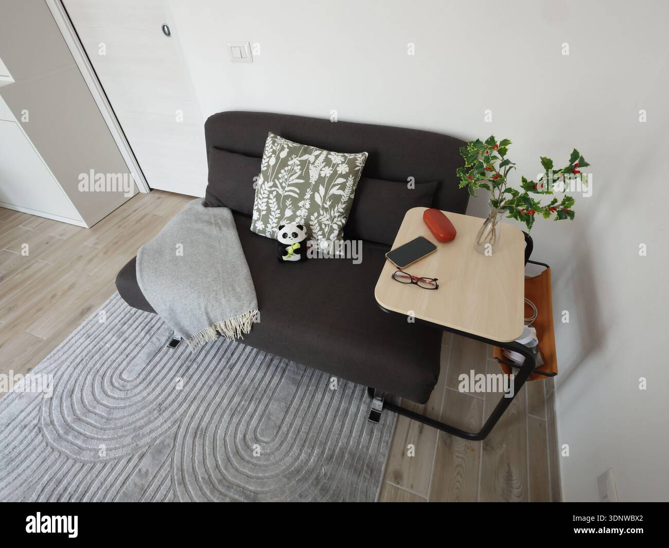 Kompaktes schwarzes ausklappbares Schlafsofa mit Überwurf und Kissen sowie Beistelltisch. Möbel für kleines Wohnzimmer. Helle Wände, grauer Teppich. Stockfoto