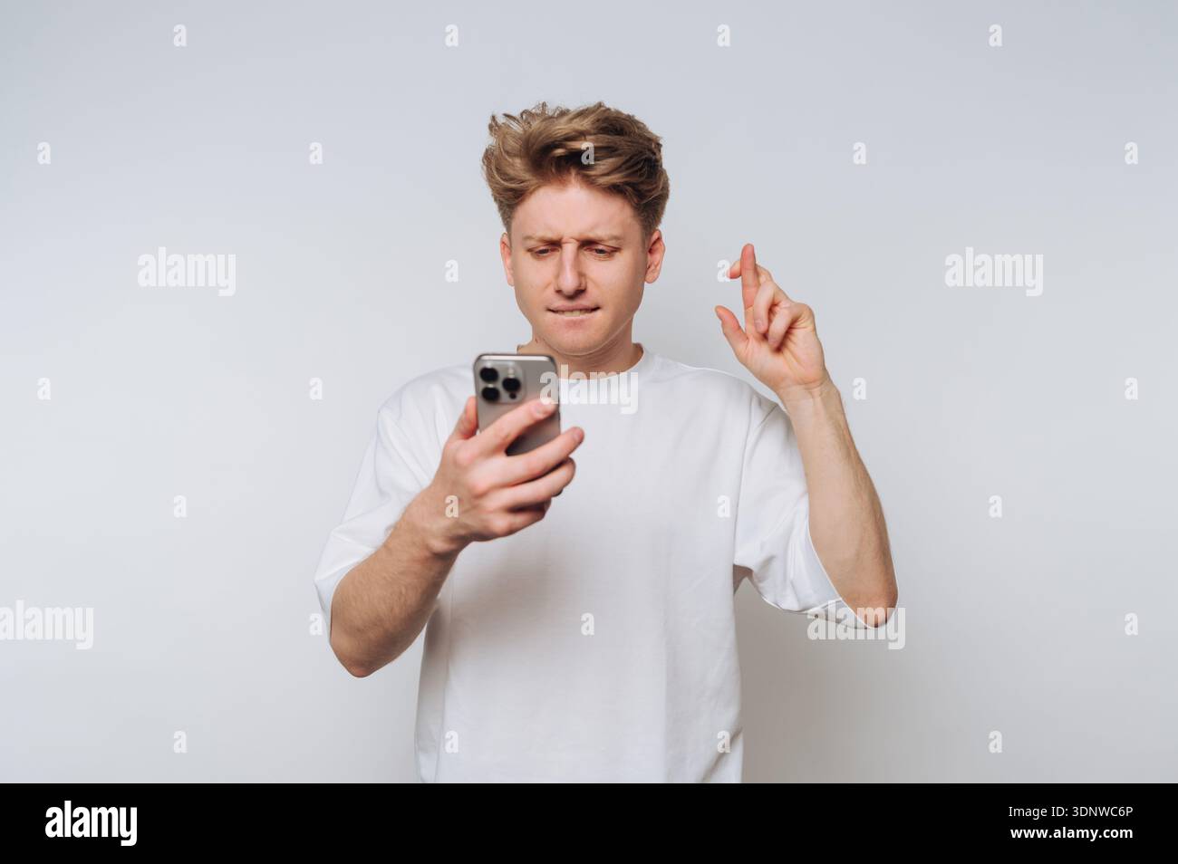 Ein junger Mann steht vor einem weißen Hintergrund und überprüft nervös sein Handy. Sein Ausdruck zeigt Vorfreude und Hoffnung, während er seinen Finger kreuzt Stockfoto