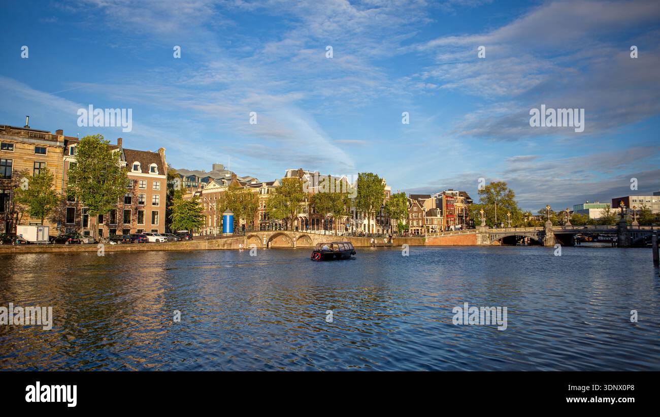 Breiter Amsterdamer Kanal mit Brücke und Skyline. Panoramablick auf den Amsterdamer Kanal mit historischen Gebäuden, Bäumen, einem kleinen Boot auf dem Wasser und einer Brücke Stockfoto