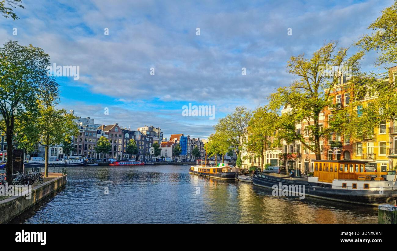 Blick auf den Amsterdamer Kanal mit Booten und Bäumen. Sonniger Morgen auf einem Amsterdamer Kanal mit historischen Gebäuden, Hausbooten, grünen Bäumen und ruhigem Wasser Stockfoto