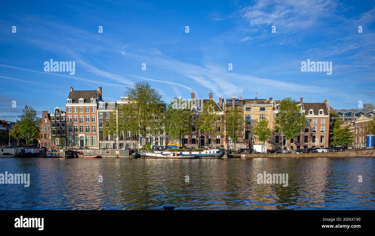 Der Amsterdamer Kanal beherbergt die Skyline über ruhiges Wasser. Weite Aussicht auf Amsterdamer Grachtenhäuser und Bäume auf dem Wasser, mit Booten, die entlang des Kais ankern Stockfoto