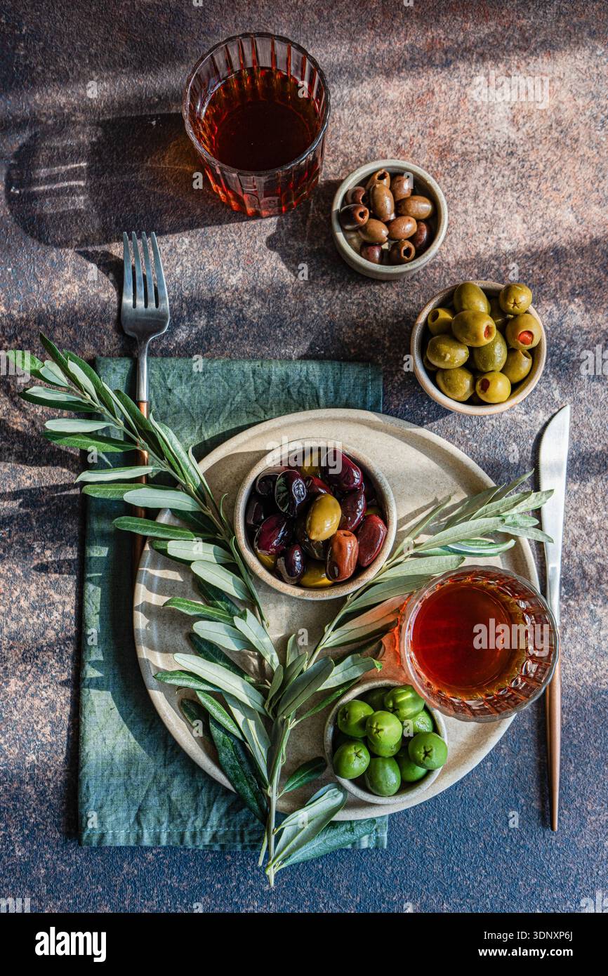 Blick von oben auf einen rustikalen Tisch mit verschiedenen griechischen Oliven und Bernsteinwein. Schüsseln mit grünen und schwarzen Oliven sind elegant auf einem Textu angeordnet Stockfoto