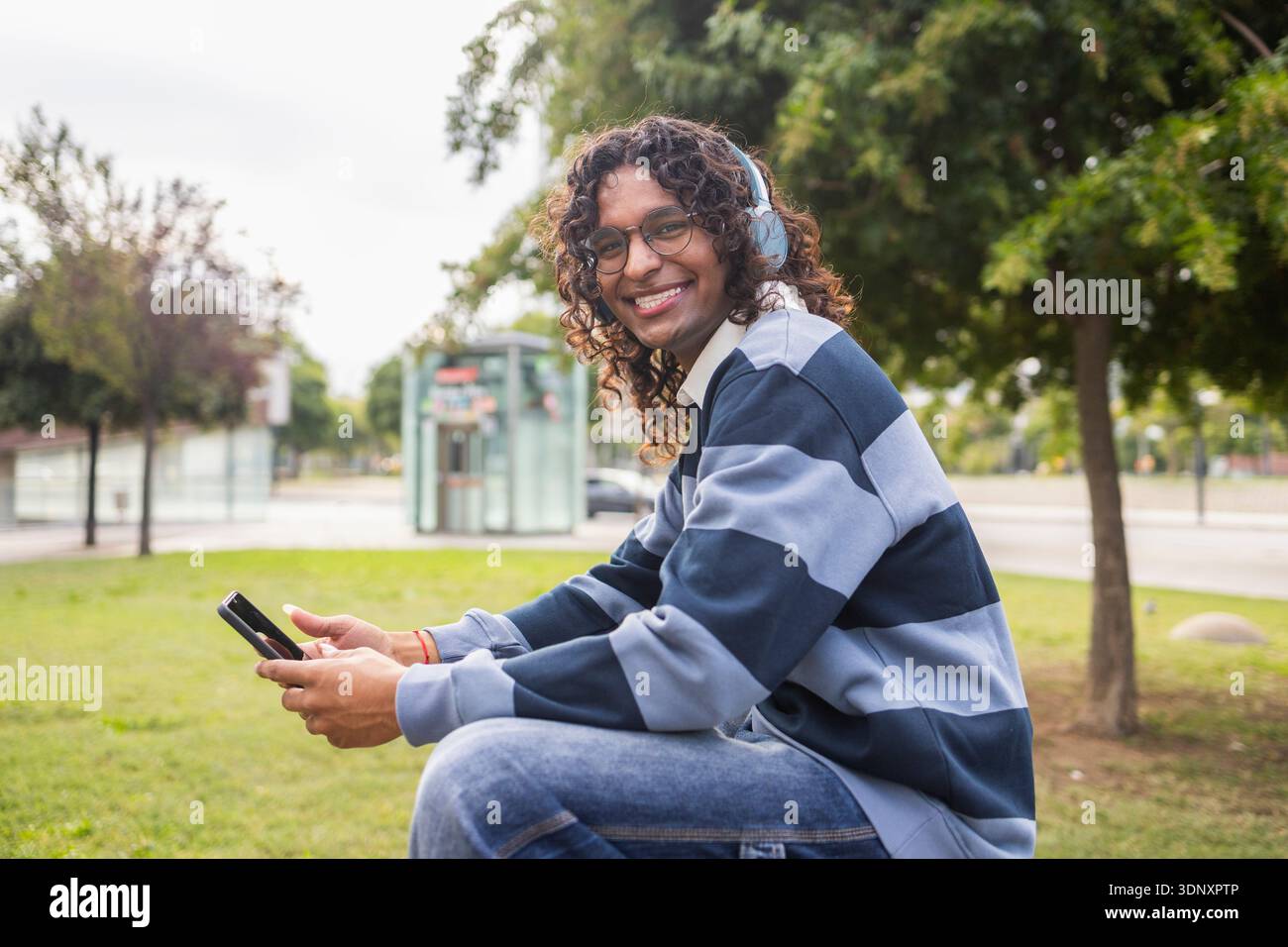 Eine freudige, nicht-binäre Person mit Kopfhörern und Telefon entspannt sich in einem Stadtpark. Diese freimütige Stadtszene mit modernem LGBT-Lifestyle ist ein Highlight für die Stadt Stockfoto
