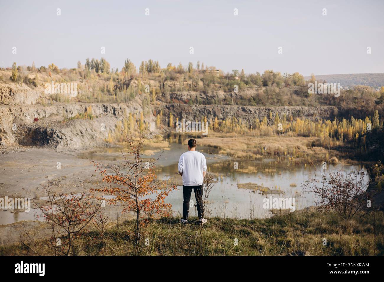 Eine Einzelfigur blickt auf einen malerischen Steinbruch, der von herbstlichem Laub umgeben ist. Das ruhige Wasser spiegelt die friedliche und introspektive Atmosphäre wider Stockfoto