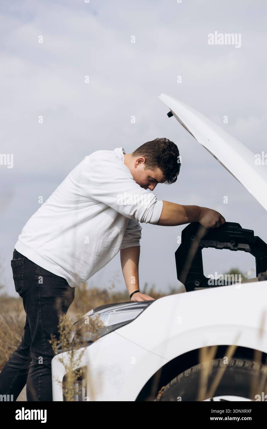 Ein junger Mann in lässiger Kleidung untersucht an einem bewölkten Tag unter der Motorhaube eines weißen Elektroautos und zeigt die Interaktion von Technologie und Stockfoto