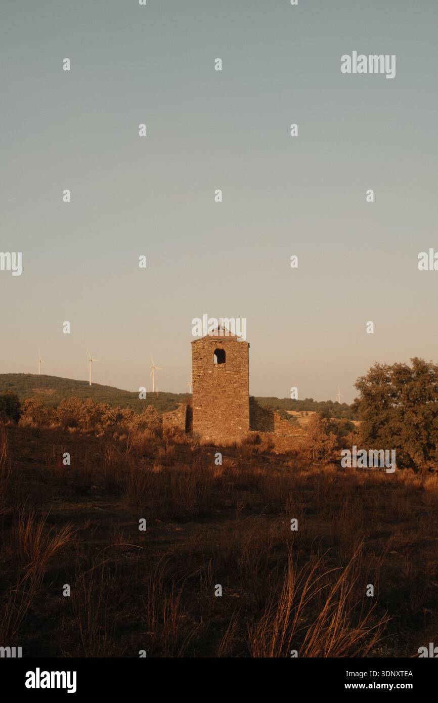 Ein einsamer Steinturm steht bei Sonnenuntergang inmitten von verbranntem Gras. In der Ferne säumen Windturbinen den Hügel, die sich mit der Silhouette des orangen Himmels verbinden Stockfoto