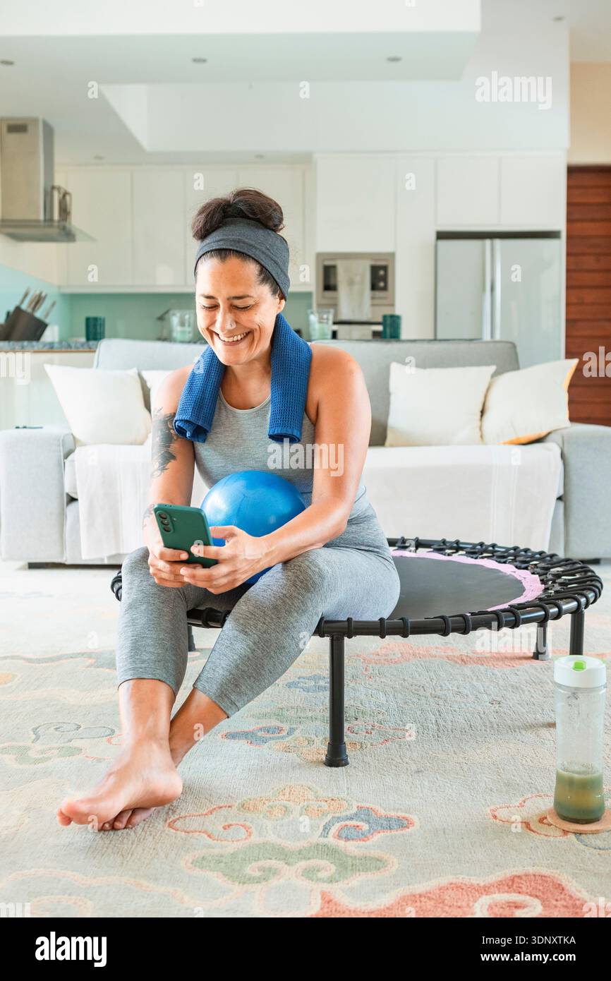 Eine fröhliche Frau macht zu Hause eine Pause auf ihrem Mini-Trampolin und hält ein Smartphone. Ein blauer Ball und eine grüne Smoothie-Flasche tragen zu ihrer gesundheitsorientierten r Stockfoto