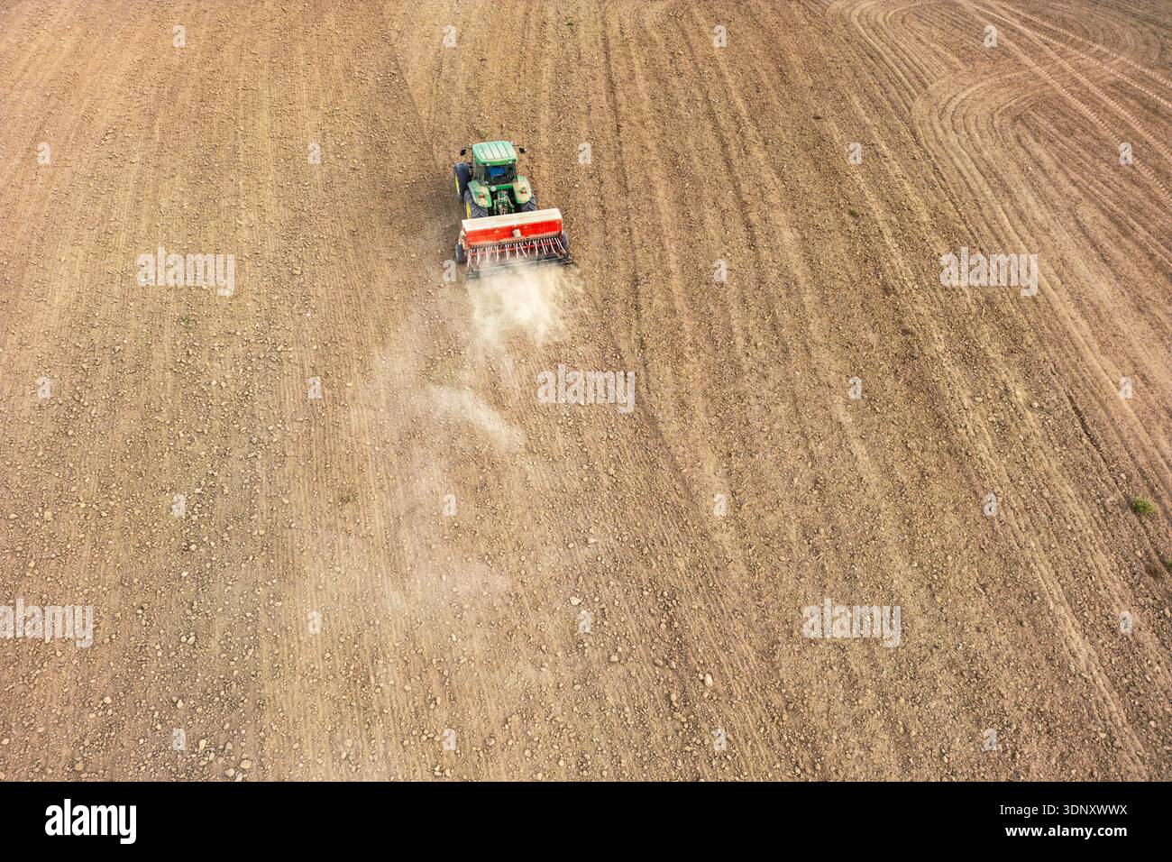 Aus der Vogelperspektive eines grünen Traktors, der von einem männlichen Landwirt angetrieben wird und ein großes, trockenes Feld pflügt. Während sich die Maschine bewegt, bläst sich Staub auf und erzeugt unterschiedliche Muster in t Stockfoto