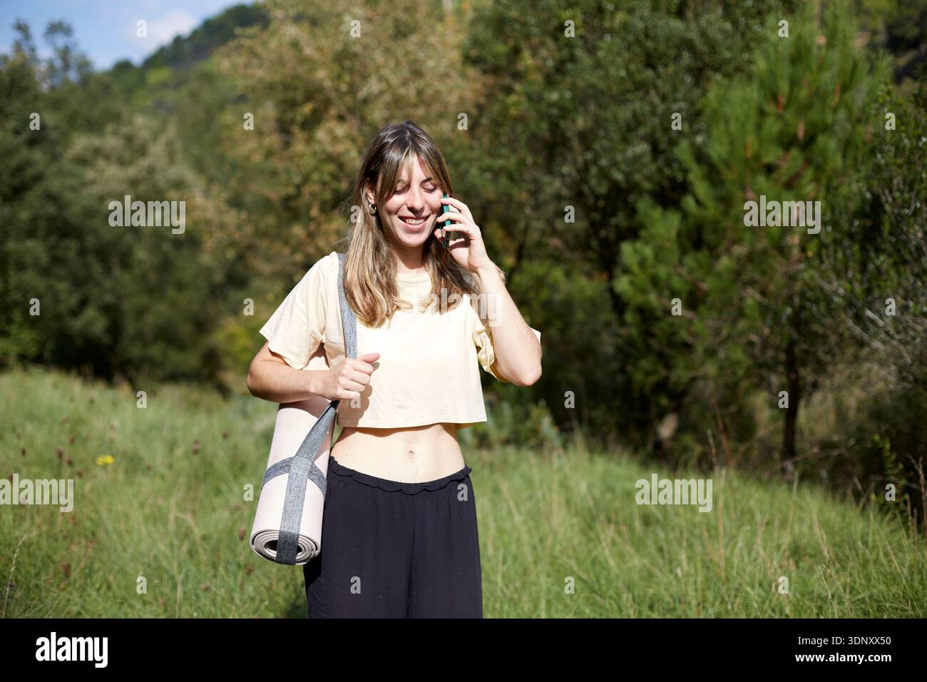 Eine Frau, die draußen steht, eine Yogamatte hält und auf einem Smartphone spricht. Sie wirkt entspannt, möglicherweise nach einer Meditationssitzung, umgeben von gre Stockfoto