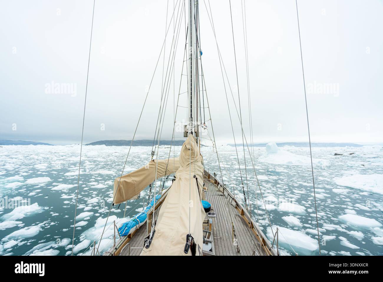 Ein Segelboot navigiert durch die eisigen Gewässer Grönlands, umgeben von schwimmenden Eisbrocken und fernen Bergen, und fängt die ruhige und doch raue Schönheit von Th ein Stockfoto