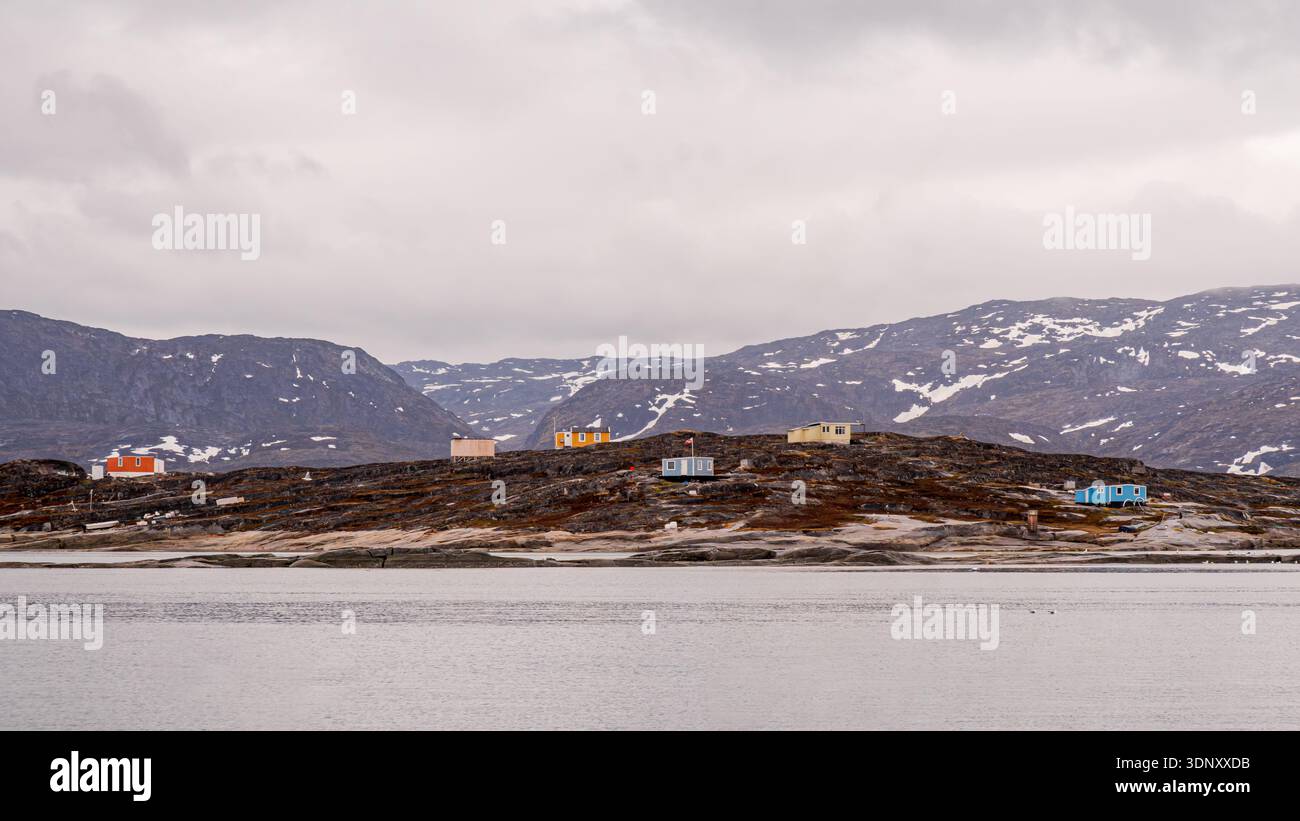 Bezaubernde farbenfrohe Hütten umgeben von dem zerklüfteten felsigen Gelände Grönlands, eingebettet unter schneebedeckten Bergen. Die ruhige und abgelegene Lage ist reflektiert Stockfoto