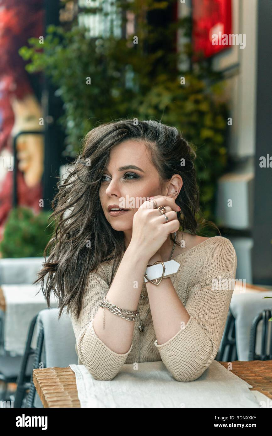 Eine Frau mit langen Haaren und elegantem Schmuck sitzt in einem stilvollen Stadtcafé. Ihr nachdenklicher Blick und ihre entspannte Haltung vermitteln ein Gefühl der Ruhe inmitten eines lebendigen Stockfoto