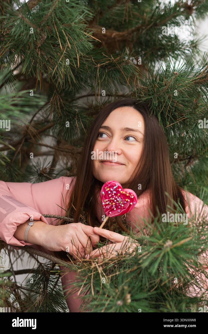 Eine Frau in rosa Jacke hält einen rosa Lutscher in Herzform und lehnt sich spielerisch an die grünen Zweige eines Weihnachtsbaums. Ihr fröhlicher Ausdruck Stockfoto