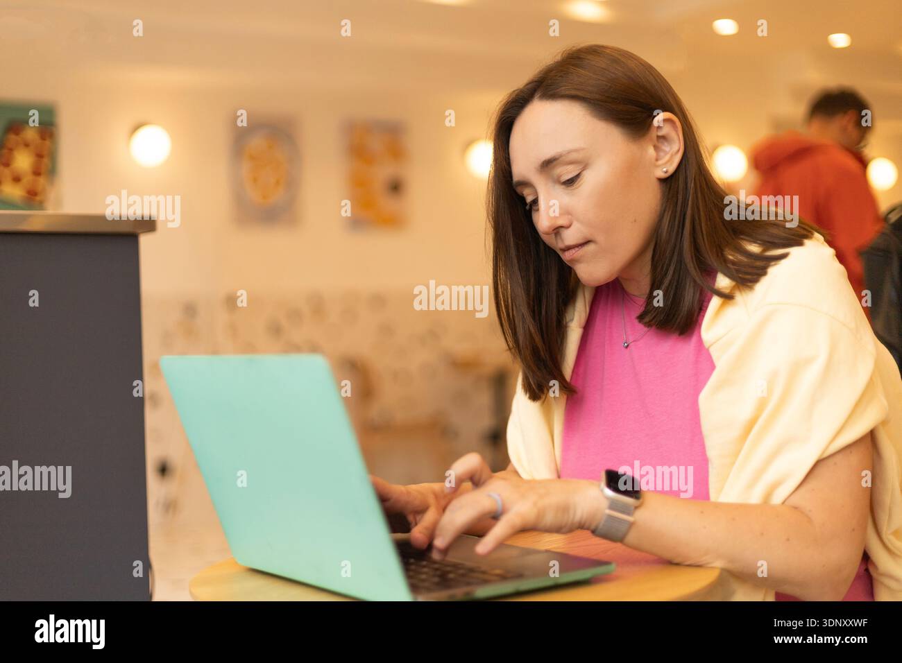 Eine junge Frau sitzt an einem Tisch in einem gemütlichen Café und tippt auf ihrem Laptop. Die entspannte Atmosphäre und die warme Beleuchtung schaffen eine perfekte Umgebung Stockfoto