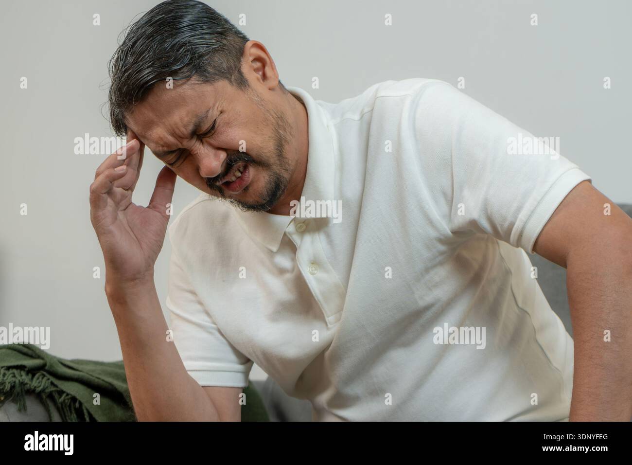 Ein junger Mann sitzt zu Hause auf einem Sofa und hält seinen Kopf vor Schmerzen, was starke Kopfschmerzen, Stress und gesundheitliche Probleme repräsentiert. Stockfoto