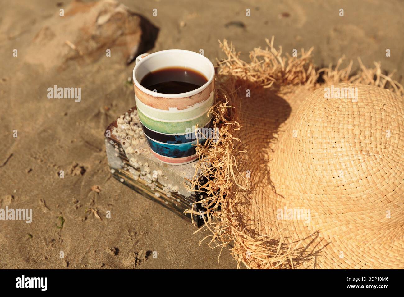 Gestreifter Becher mit Kaffee und Strohhut auf Sand am Meer Stockfoto
