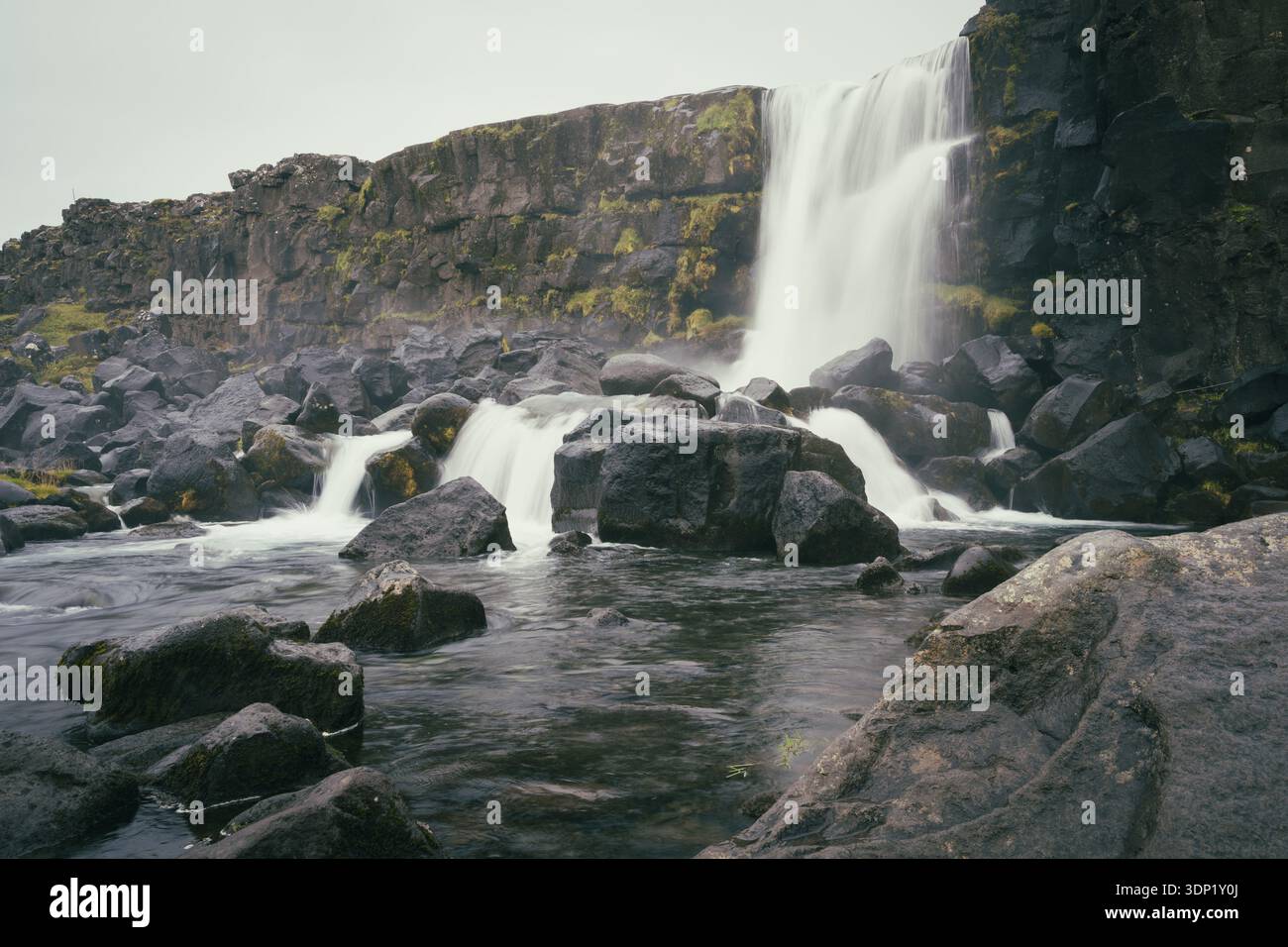 Wasserfall über dunkle Vulkanfelsen im Thingvellir Nationalpark unter einem bewölkten isländischen Himmel Stockfoto