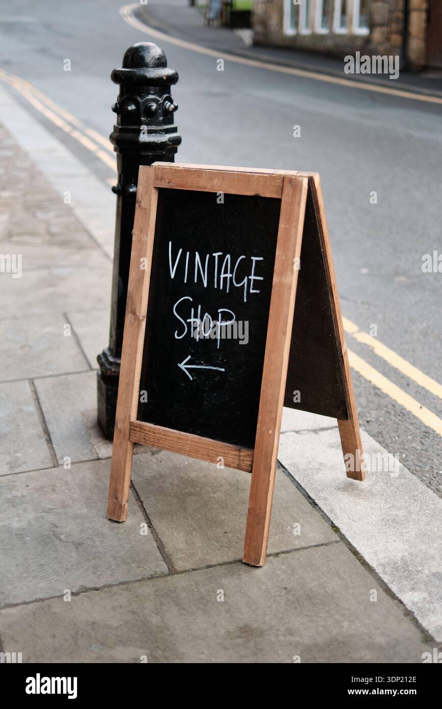 Hölzerne Kreidetafel mit A-Rahmen, die einen Vintage-Shop mit einem Pfeil nach links auf einem mit Steinmarkierungen versehenen Bürgersteig in Hebden Bridge, Yorkshire, bewirbt. Stockfoto
