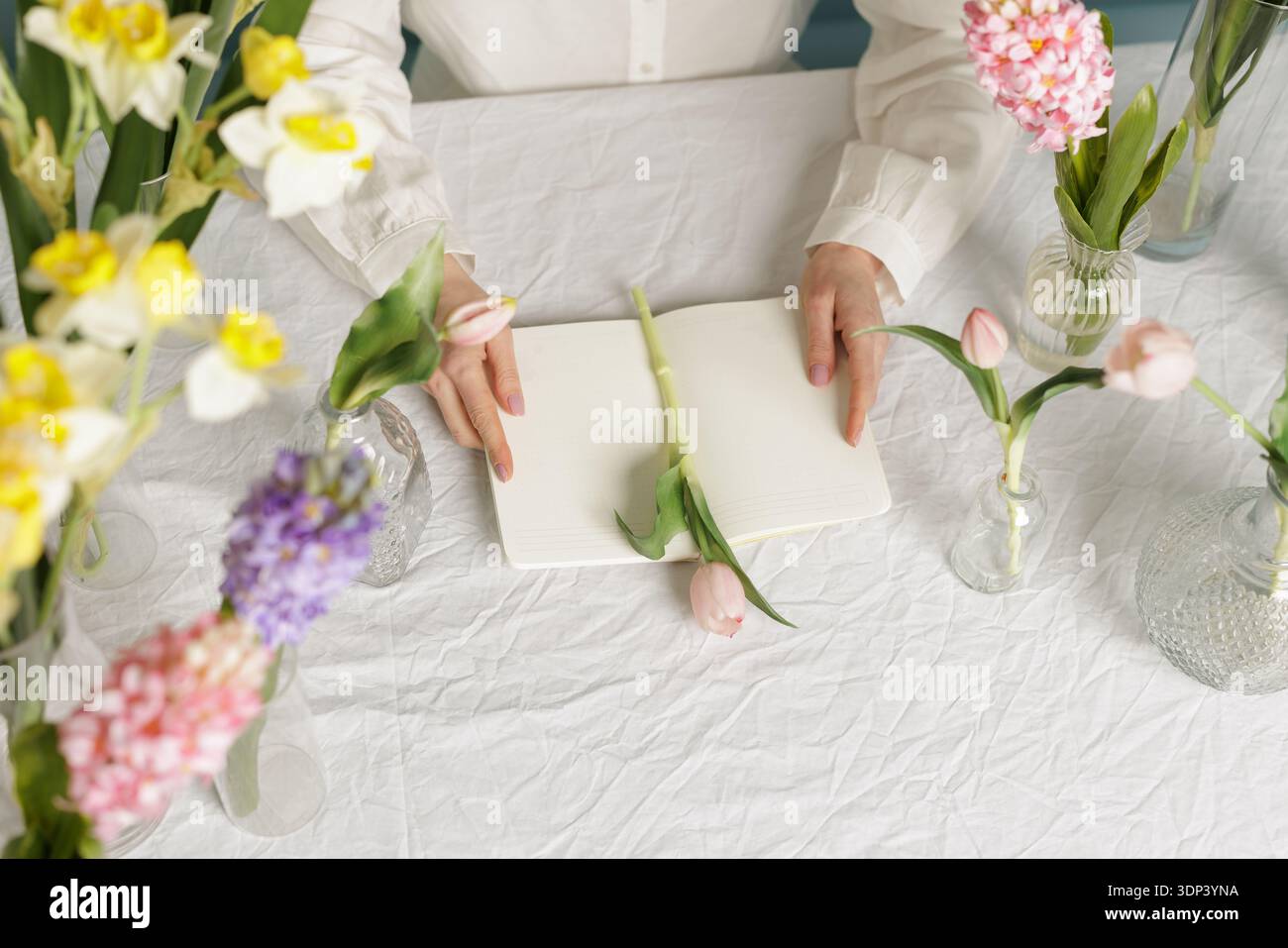 Draufsicht auf den Blumenarbeitsplatz mit offenem Notizbuch und Frühlingsblumen in Vasen. Ideal für Floristik-Branding, saisonales Marketing und kreative Geschäfte Stockfoto