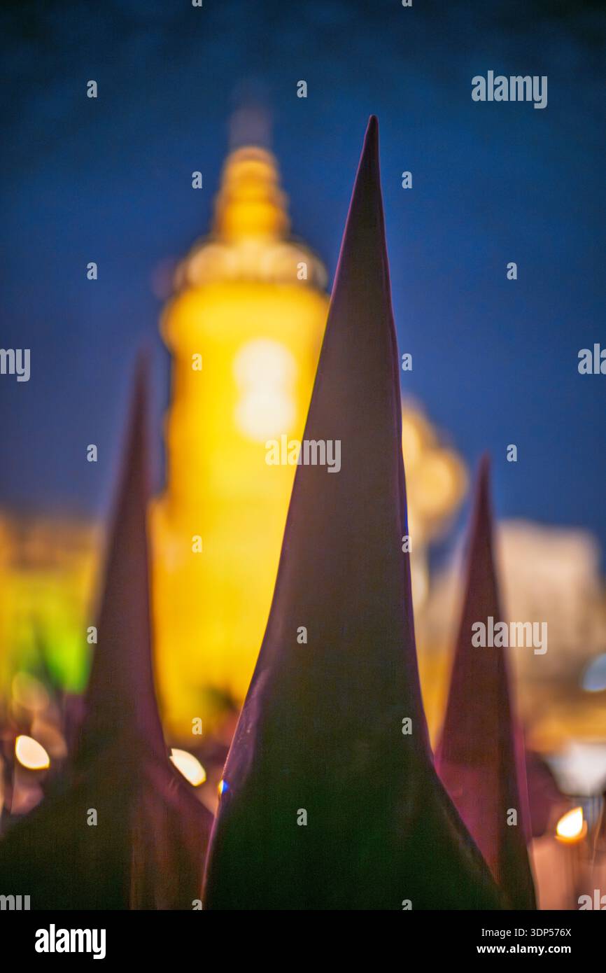 Die Nazarener der Bruderschaft La Estrella ziehen nachts während der Karwoche in Sevilla, Spanien. Konische Hauben im Vordergrund verschwimmen die Carmen Chapel. Stockfoto