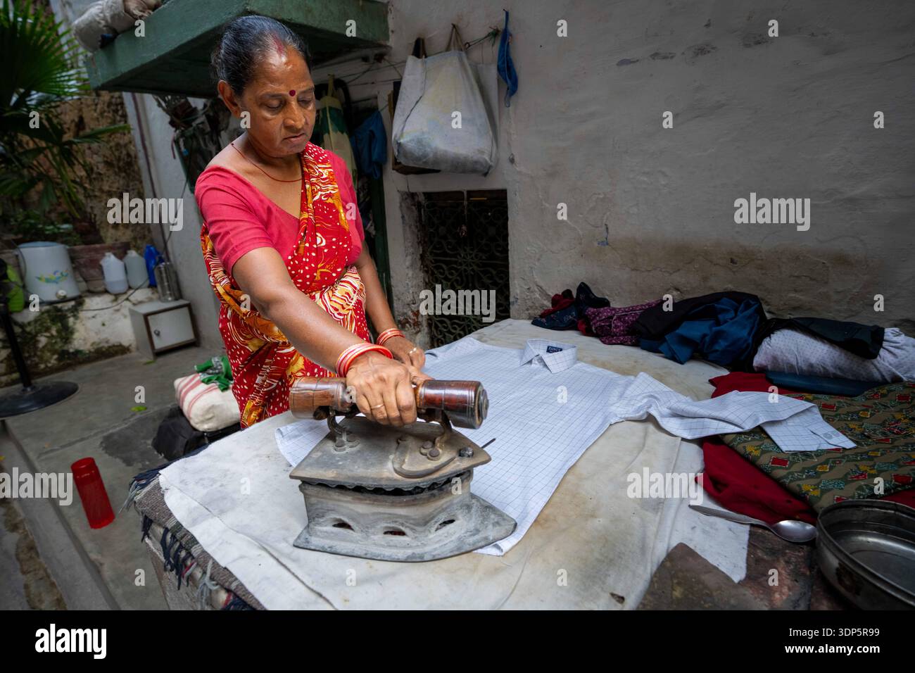 Die indische Dame in rotem Sari benutzt auf der Straße ein dickes, schweres Eisen, um Hemd zu bügeln Stockfoto