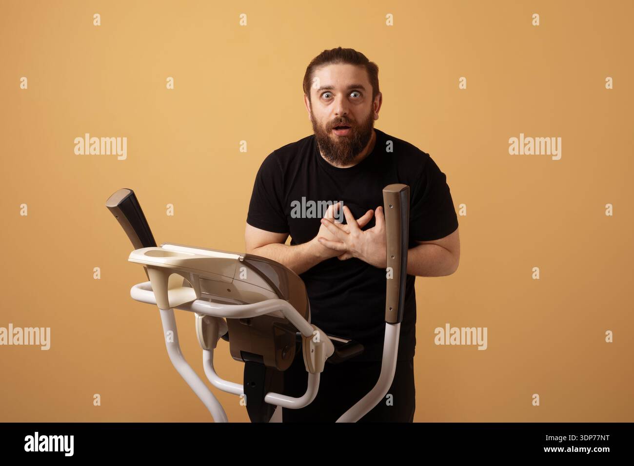 Bärtiger Mann in schwarzem T-Shirt, der überrascht aussieht, während er auf dem Heimtrainer vor beigefarbenem Hintergrund steht. Stockfoto