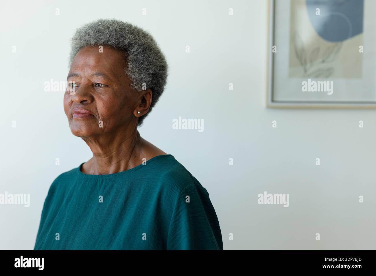 Ältere afrikanische Frau, die im schlichten Zimmer mit blaugrünen Nieten und gerahmten Kunstwerken, Kopierraum, zurückblickt Stockfoto